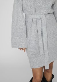 Robe en tricot gris clair avec une taille ceinturée, des manches larges et un tissu texturé. Présente un ourlet brut avec une finition douce.