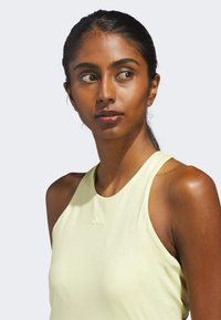 Vaaleankeltainen ilman hihoja oleva urheiluyläosa, jossa on pyöreä pääntie ja kohokuvioitu logo. Silkkinen, joustava kangas, sopiva muoto.