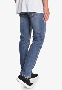 Jeans in denim blu da uomo con una vestibilità slim, lavaggio sbiadito e tasche posteriori. Indossati con sneaker nere, dotate di suole bianche.