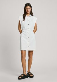 Witte, kortarmige shirtjurk met een knoopsluiting aan de voorkant, een getailleerde taille en een rechte zoom. Gecombineerd met zwarte gespte sandalen.