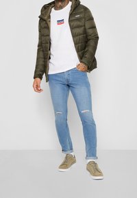 Olivgrön puffjacka, vit t-shirt med Levi's-logga, ljusblå slitna jeans och bruna sneakers med vita sulor.