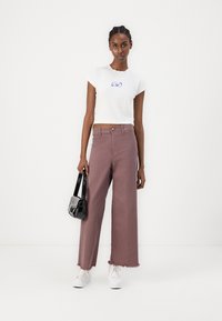White cropped t-shirt with purple embroidery, paired with high-waisted, wide-leg mauve jeans featuring frayed hems. Black handbag and white sneakers.