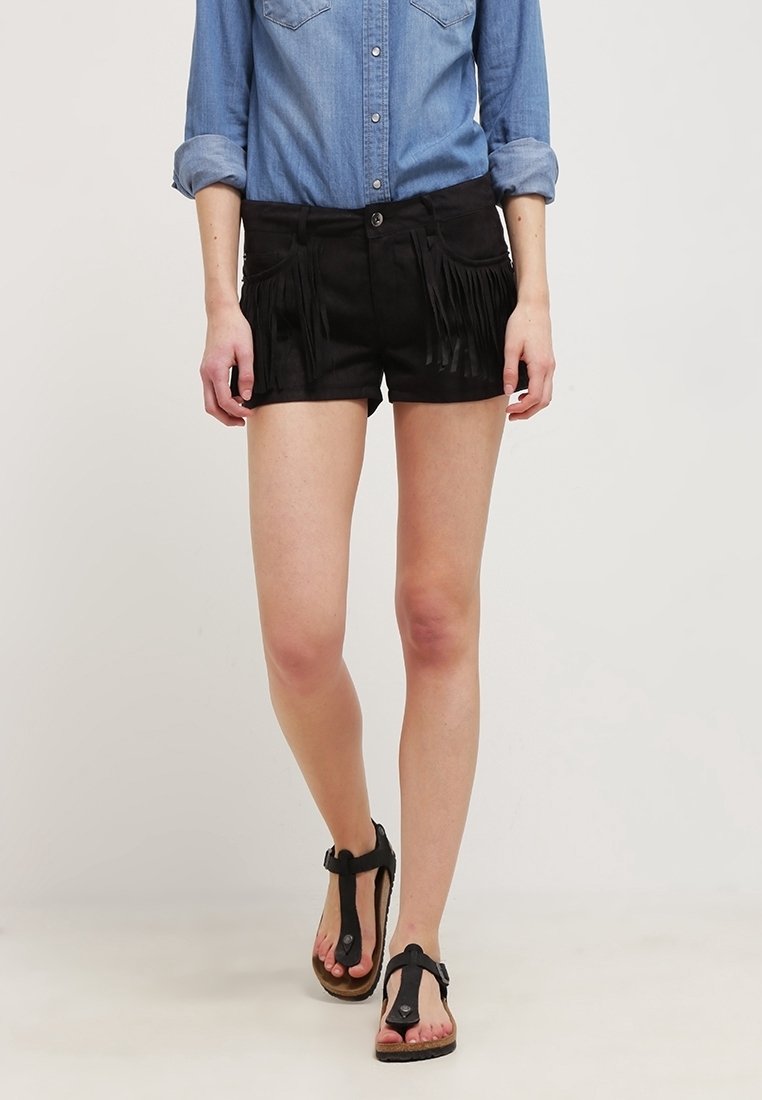 Personne portant une chemise bleue aux manches retroussées, un short noir à franges et des sandales noires, debout sur un fond clair.