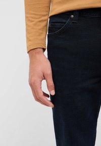 Dunkelblaue Denim-Jeans mit schmaler Passform, mit einer Vorderseite-Tasche und Metallknopf-Details, getragen mit einem senffarbenen Langarmshirt.