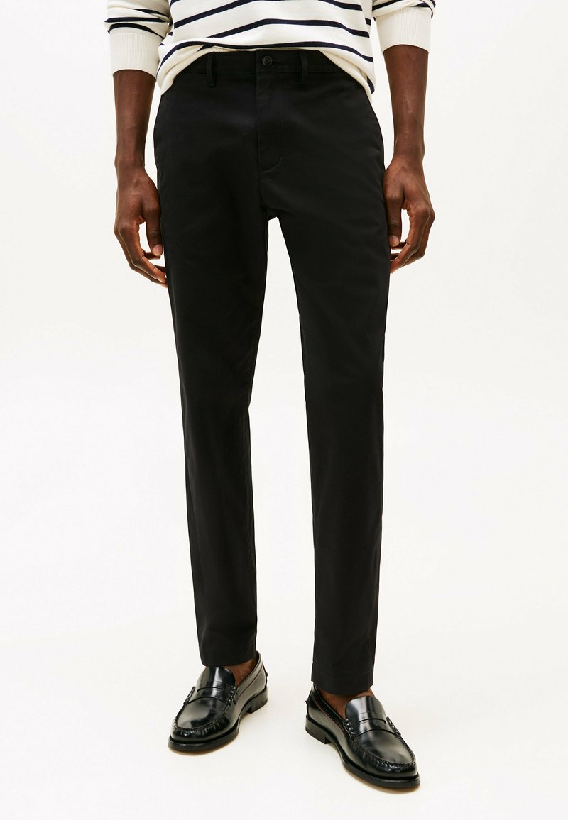 Pantaloni neri slim fit con una texture liscia, tasche anteriori e chiusura con bottone, abbinati a mocassini neri.