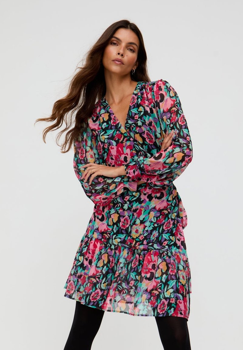 Frau mit langen Haaren, die ein buntes, geblümtes Kleid mit transparenten Puffärmeln trägt und mit verschränkten Armen vor einem einfarbigen Hintergrund posiert.