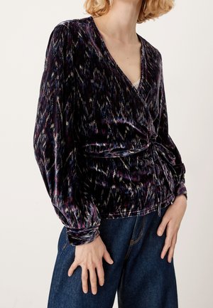 Mujer con blusa cruzada de estampado oscuro con mangas largas abullonadas y vaqueros azules, manos relajadas a los lados.