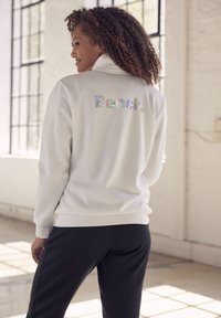 Weißer Sweatshirt mit hohem Kragen, mit schimmerndem "Bench."-Text auf dem Rücken. Kombiniert mit dunklen Hosen in einem hellen Innenraum.