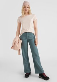 Lichtroze katoenen T-shirt, teal wijde broek, beige gebreide trui over de arm, en zwarte wandelschoenen met rode accenten.