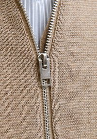Tuvumā redzama sudraba rāvējslēdzis, daļ�ēji atvērts uz beige adītā džempera, atklājot zem tā zilu un balto svītrainu krekliņu.