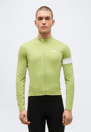 Moški, ki nosi svetlo zeleno kolesarsko majico z dolgimi rokavi z belim trakom na levi roki in črne kolesarske hlače, stoji obrnjen naprej.