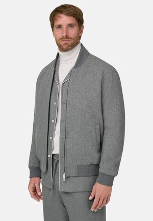 Homme portant une veste grise zippée sur une chemise grise boutonnée et un col roulé blanc, debout devant un fond clair uni.