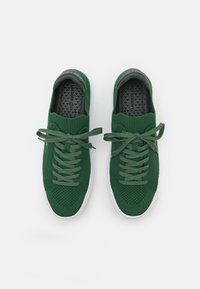 Sapatilhas verdes de malha com textura em rede, com atacadores planos e uma sola branca de borracha. Apresentam uma palmilha com marca e um design de bico arredondado.