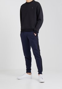 Sudadera negra con puños texturizados y un pequeño logo; pantalones de chándal azul marino con rayas laterales; zapatillas blancas. Conjunto de ropa deportiva informal.
