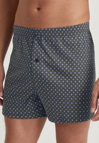 Un caleçon en tissu bleu foncé avec un motif géométrique de petites croix vertes et blanches ; terminé par un bouton à l'avant et une taille élastique.