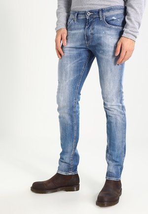 Man draagt slank gescheurde lichtblauwe spijkerbroek, grijs shirt met lange mouwen en donkerbruine leren laarzen, staand op een witte achtergrond.