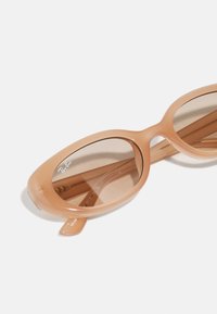 Ochelari de soare cu nuanță maronie, cu un cadru din plastic neted, de culoare piersică deschisă. Lentile ovale și un design subțire, curbat; prezintă un logo Ray-Ban.