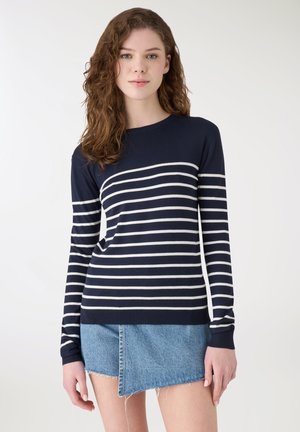 Giovane donna con capelli castani ondulati che indossa un maglione a maniche lunghe a righe blu navy e bianche e una mini gonna di jeans asimmetrica.