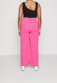 Person som vänder ryggen till, iklädd en svart asymmetrisk linne, ljusrosa vida byxor och vita sportskor mot en neutral bakgrund.