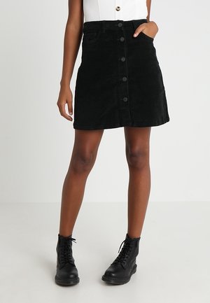 Femme porte une jupe en velours côtelé noire boutonnée devant, un haut blanc et des bottines noires à lacets, debout avec une main dans la poche.