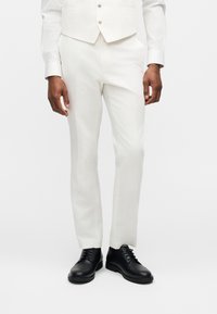 Άνδρας που φορά άσπρο παντελόνι φόρμας, λευκό γιλέκο και μαύρα παπούτσια φόρμας με κορδόνια, στέκεται μπροστά σε απλό ανοιχτό φόντο.