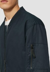 Moški nosi temno modro bomber jakno z zadrgo na levi rukavu, stoji pred enobarvno svetlo ozadje.