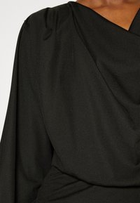 Gros plan sur une personne portant un haut en tissu noir drapé avec un motif texturé subtil et un détail froncé à l'épaule.