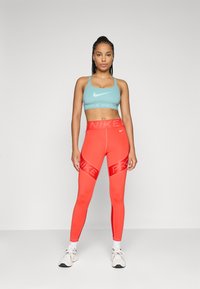 Žena stoji u cijan sportskom grudnjaku i svijetlosnim crvenim Nike Pro tajicama, s bijelim tenisicama, na jednobojnoj sivoj pozadini.