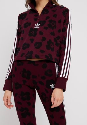 Kobieta w bordowym dresie Adidas z czarnym kwiatowym wzorem i białymi paskami na rękawach i nogawkach, logo Adidas widoczne na piersi i udzie.