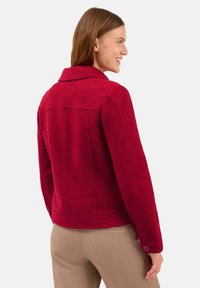Femme portant une veste texturée rouge ajustée avec un col et des poignets boutonnés, assortie à un pantalon beige, de profil en souriant.