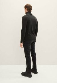 Schwarzer Rollkragenpullover, schmale Passform, gerippter Kragen und Bündchen, kombiniert mit dunklen Jeans und schwarzen Schuhen, von hinten vor einem neutralen Hintergrund gezeigt.