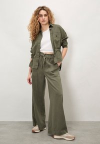 Camisa corta de botón de color verde oliva, ligera, con mangas remangadas, combinada con pantalones anchos sueltos a juego y zapatillas beige.