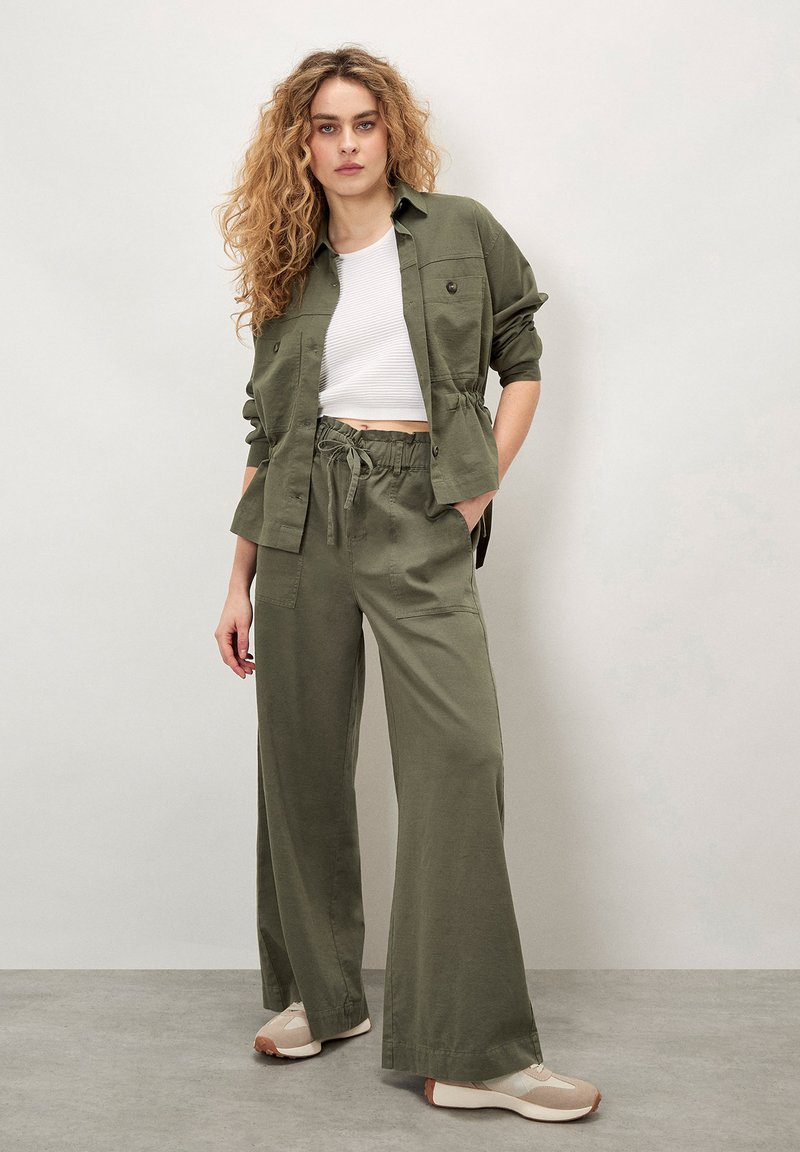 Camisa corta de botón de color verde oliva, ligera, con mangas remangadas, combinada con pantalones anchos sueltos a juego y zapatillas beige.