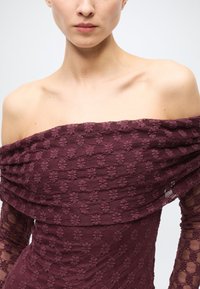 Off-the-shoulder burgundy dress with textured floral pattern, layered neckline, and sheer lace long sleeves. Fitted bodice design.