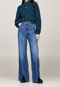 Vrouw in een teal gesprenkeld gebreid sweater, ingestopt in hoge, wijde blauwe jeans met voorknipsels, gedragen met zwarte schoenen.