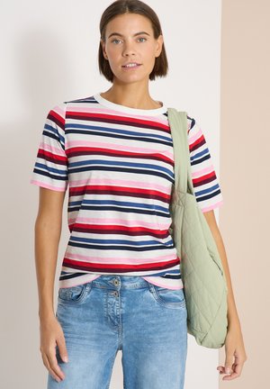 Frau trägt ein kurzärmeliges gestreiftes Hemd in Rot, Marineblau, Weiß und Rosa mit hellblauen Jeans und hält eine hellgrüne gesteppte Tragetasche.