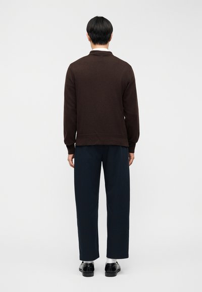 Καφέ πουλόβερ με υφή, μακριά μανίκια και κολάρο, συνδυασμένο με μπλε σκούρο παντελόνι. Ορατές ραφές στη βάση του πουλόβερ.