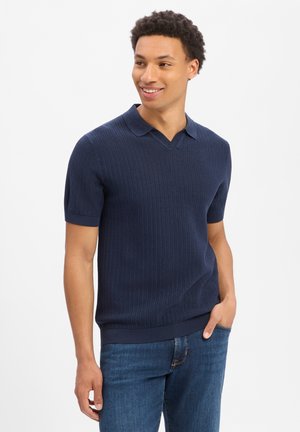 Junger Mann lächelnd, trägt ein marineblaues kurzärmeliges Strick-Poloshirt und blaue Jeans, steht mit einer Hand in der Tasche vor weißem Hintergrund.