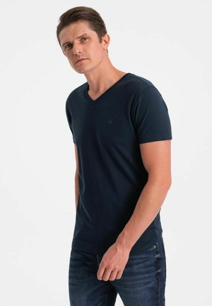 Navyblaues V-Ausschnitt-T-Shirt aus weichem Stoff, mit kurzen Ärmeln und einem dezenten Logo auf der Brust. Kombiniert mit dunklen Denim-Jeans.