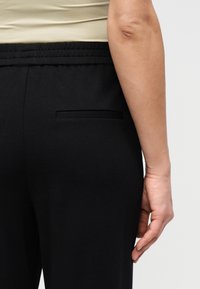 Πλάγια όψη δεξιά ενός ατόμου που φοράει μαύρο παντελόνι με ελαστική μέση και πίσω τσέπη με επένδυση, το χέρι χαλαρό δίπλα στη λεκάνη.