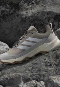 Chaussure de randonnée beige et blanche avec trois bandes blanches, semelle texturée et languette de traction reposant sur des rochers gris rugueux.