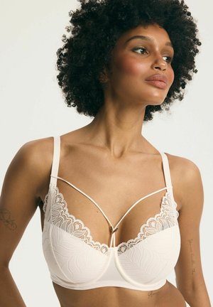 Kobieta z kręconymi włosami nosi biały koronkowy biustonosz z cienkimi ozdobnymi ramiączkami na gładkim jasnym tle.
