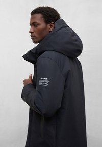 Czarna ocieplana kurtka z wysokim kołnierzem i dużym kapturem. Na górnej części rękawa znajduje się naszywka z logo i napisem. Gładka, matowa faktura materiału.