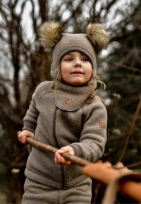 Beige fleeceoverall med dragkedja framtill, hög krage och en etikett. Hatten har två fuskpälsbollar på toppen. Naturlig utomhusmiljö.
