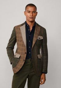 Bruin en groen geruit blazer met een contrasterende grijze revers, bruine knopen en opgestikte zakken; gedragen over een marineblauw shirt en donkere broek.