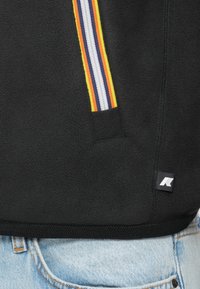 Giacca in pile nera con tasca con zip a strisce multicolori e piccolo etichetta in tessuto, indossata sopra jeans di denim azzurri.
