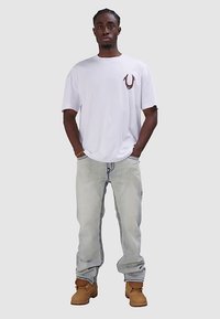 Biała koszulka z krótkim rękawem z bordowym logo na piersi, zestawiona z jasnymi dżinsami i brązowymi butami roboczymi.