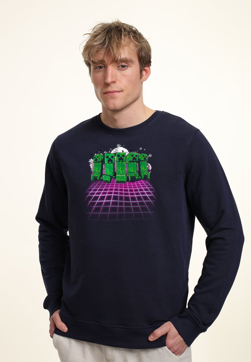 Felpa blu navy con una grafica di personaggi pixelati verdi su uno sfondo a griglia viola. Tessuto morbido e maniche lunghe con polsini a coste.