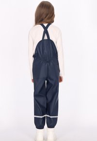 Vaikas, dėvintis tamsiai mėlynas vandeniui atsparias kelnes su kryžminėmis petnešomis ir baltus batus, stovi atsukęs nugarą prieš paprastą baltą fono.