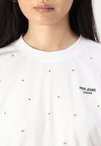 Balta medvilninė marškinėlė su išsibarsčiusiais juodais ir sidabriniais aksominiais akmenukais bei šonu nespalvotu logotipu "PEPE JEANS LONDON".